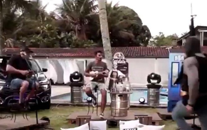 O grupo de pagode Aglomerou toca, enquanto um policial aparece com uma arma no meio da live
