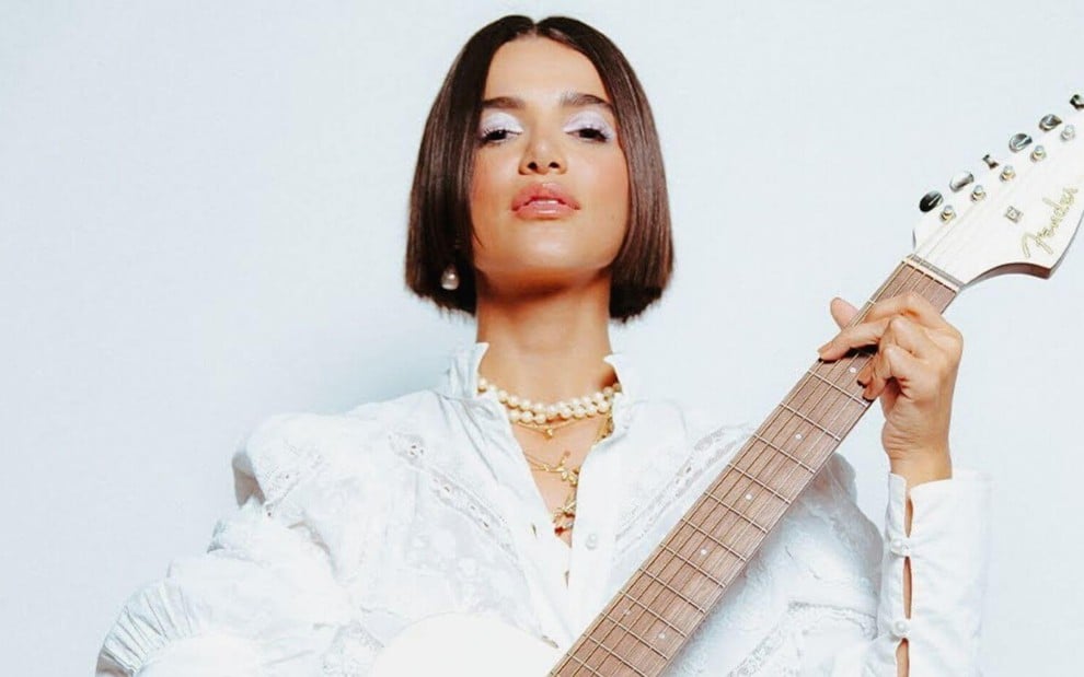 Toda de branco, a cantora Manu Gavassi segura uma guitarra e faz pose para a câmera