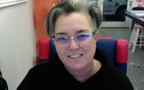 Sentada de frente para a câmera de um computador, Rosie O'Donnell sorri, de óculos e cabelo branco
