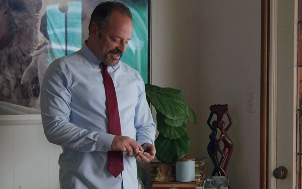 Usando camisa de manga longa e gravata, Gil Bellows usa álcool gel na mão esquerda em em cena da minissérie Love in the Time of Corona