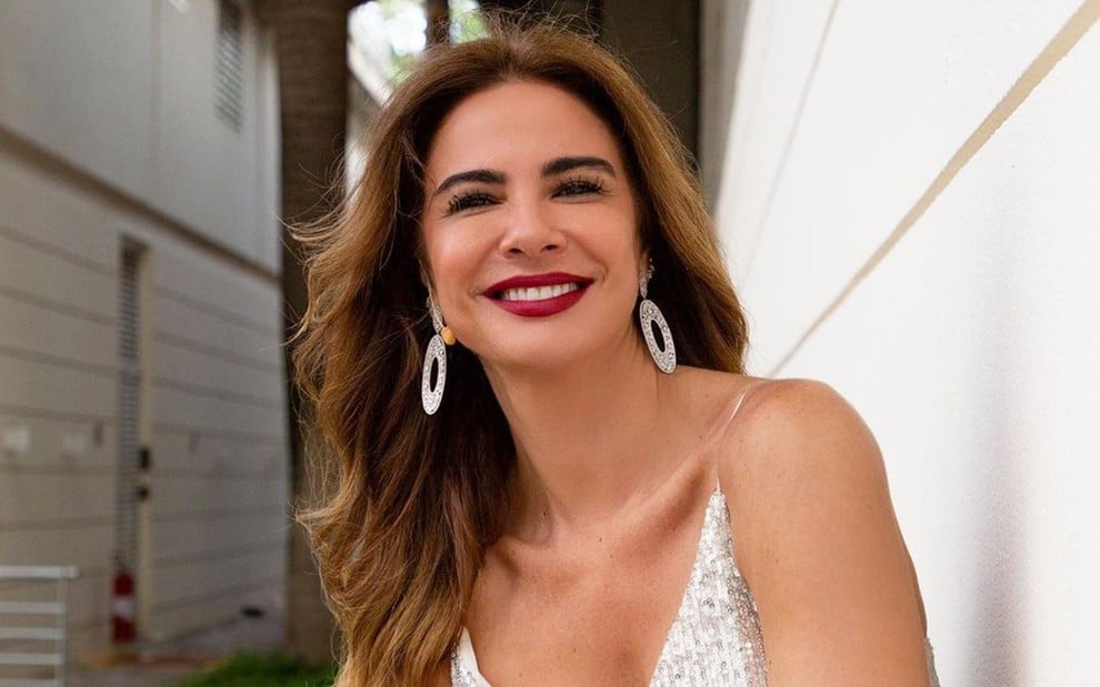 Luciana Gimenez posa para foto, ela olha para a câmera e sorri; a apresentadora está de cabelo solto, brincos pratas e vestido prata com brilhos