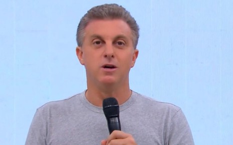 O apresentador Luciano Huck segurando microfone em frente a um fundo branco