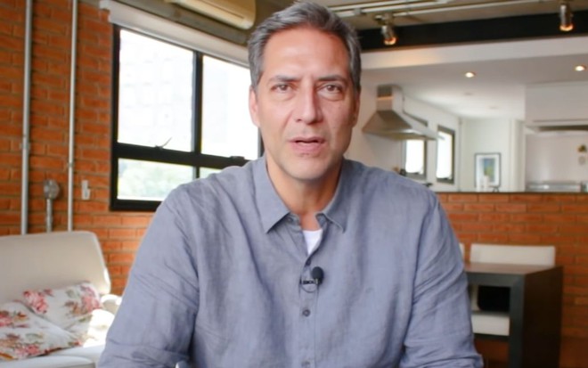 Imagem de Luís Ernesto Lacombe em sua casa, usando uma camisa azul