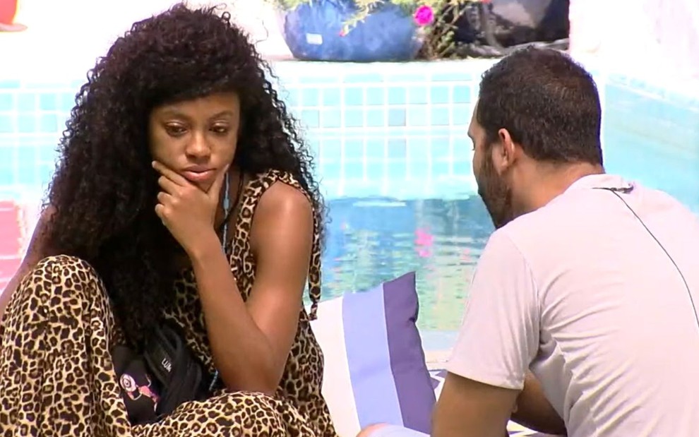 Lumena Aleluia com a mão no queixo, pensativa, em conversa com Gilberto Nogueira, de costas, sentados perto da piscina