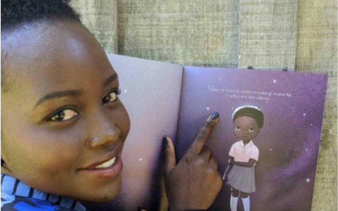 Lupita Nyong'o aponta para gravura do livro Sulwe, escrito pela atriz, enquanto olha em direção à câmera
