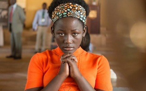 A atriz Madina Nalwanga usa uma blusa laranja e está com as duas mãos no queixo em cena como Phiona Mutesi no filme Rainha de Katwe (2016)
