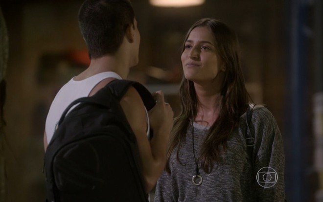 De costas, Arthur Aguiar grava cena de Malhação, usando regata branca e mochila nas costas, ao lado de Maria Joana, com cabelo solto e blusa lisa