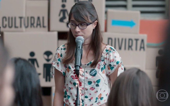 A atriz Daphne Bozaski em frenta a um microfone, com expressão envergonhada, em cena de Malhação