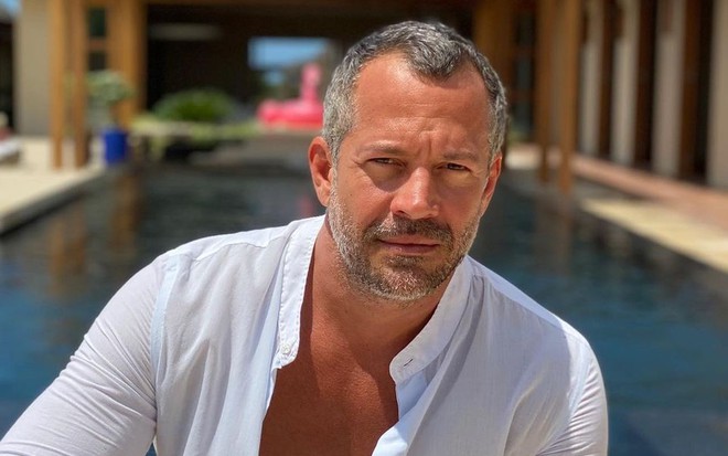 Malvino Salvador sentado em frente à piscina, de camisa branca com o peitoral à mostra
