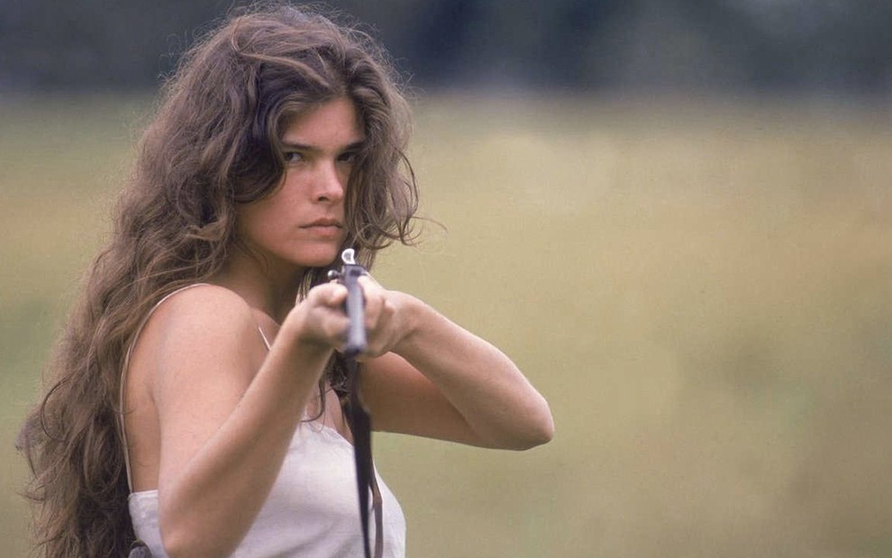 A atriz Cristiana Oliveira com os cabelos desgrenhados como Juma aponta uma arma em direção à câmera em cena de Pantanal