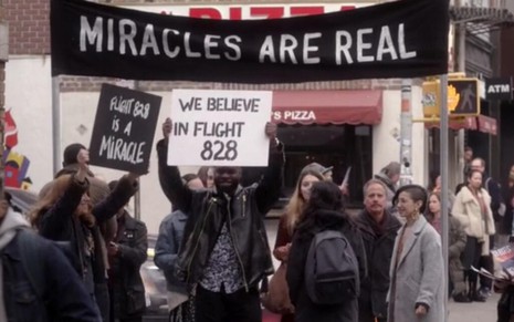 Várias pessoas no meio de uma calçada erguem cartazes e faixas com dizeres religiosos na série Manifest