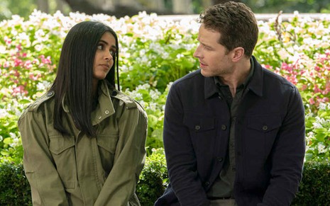Sentados em um banco, com flores atrás, Parveen Kaur e Josh Dallas trocam olhares na série Manifest