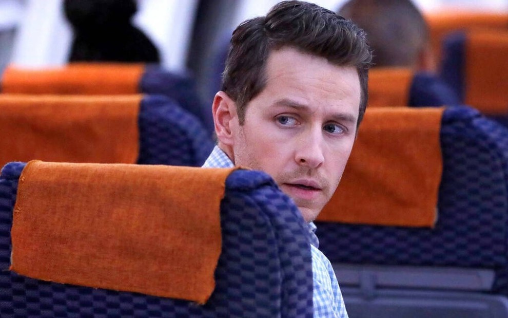 Dentro de um avião, sentado em uma poltrona azul escura, o ator Josh Dallas olha para trás em cena da série Manifest
