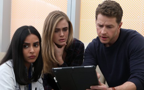 Parveen Kaur olha para o nada enquanto Melissa Roxburgh e Josh Dallas, concentrados, encaram um tablet na série Manifest