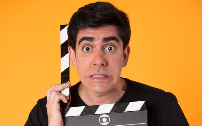 Marcelo Adnet segura claquete e faz cara esquisita. Fundo laranja