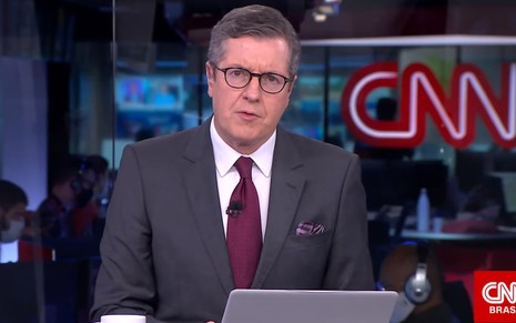 Márcio Gomes com um terno cinza, camisa branca e gravata vinho, sentado na bancada de um telejornal da CNN Brasil
