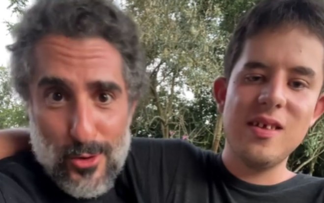 Marcos Mion ao lado de seu filho, Romeo Mion, em local a céu aberto com árvores, em frame de vídeo