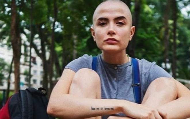 Maria Casadevall sentada no chão, com as pernas encolhidas, camiseta azul com suspensório e a cabeça raspada
