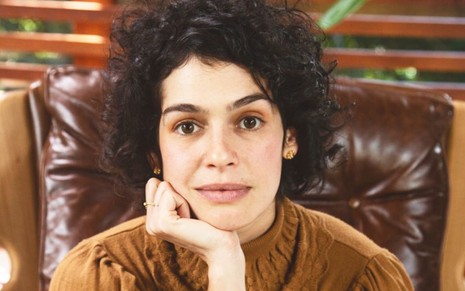 Maria Flor está sentada em uma poltrona marrom; ela usa blusa gola alta marrom e está com a mão no queixo