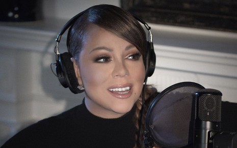 A cantora Mariah Carey com um fone de ouvido do tipo headseat, ela está de preto em frente a um microfone de formato redondo