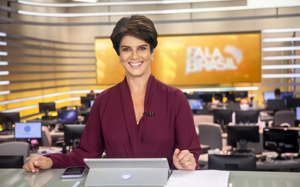 A apresentadora Mariana Godoy sorri com os dois braços sobre a mesa no estúdio do Fala Brasil, da Record