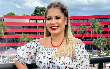 Marília Mendonça está em frente a um prédio vermelho e posa para foto; ela usa vestido branco com detalhes coloridos, colar vermelho no pescoço e está de cabelos solto