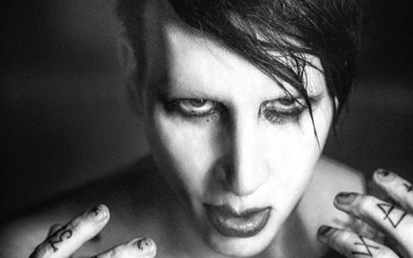 Foto em preto e branco de Marilyn Manson maquiado, com franja lateral e as mãos tatuadas na altura do rosto