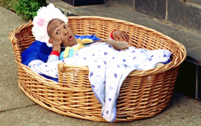 Rosto do ator Marlon Wayans no corpo de um bebê, deitado dentro de uma cesta, com a boca aberta
