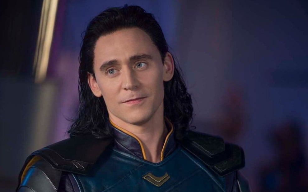 Ator Tom Hiddleston com cabelos longos e roupa em tons azulados do personagem Loki em cena de Thor: Ragnarok (2017)