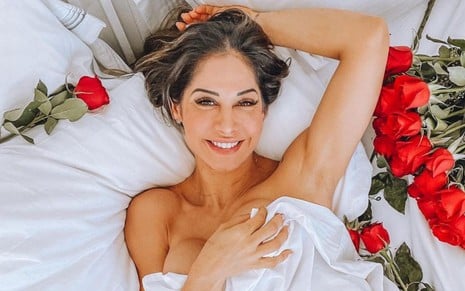 Mayra Cardi deitada em uma cama com lençóis brancos; ela sorri e posa ao lado de rosas vermelhas
