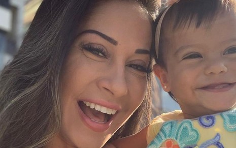 Mayra Cardi posa sorridente com a filha, Sophia, que está usando uma tiara de flor laranja e uma roupinha colorida