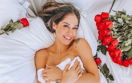 Mayra Cardi deitada em uma cama com lençóis brancos; ela sorri e posa ao lado de rosas vermelhas