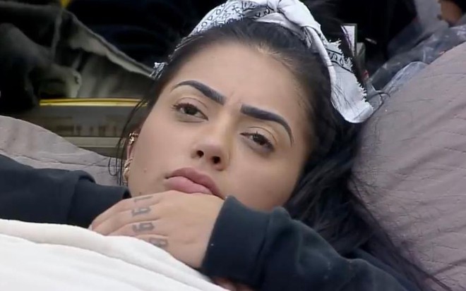 MC Mirella aparece deitada na cama, com uma coberta, blusa de manga preta e lenço branco no cabelo, pensativa
