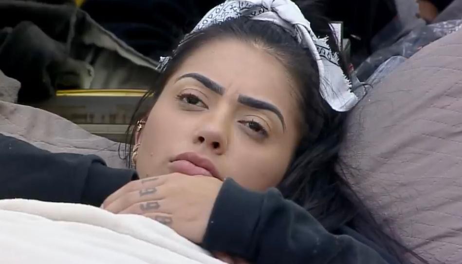 MC Mirella aparece deitada na cama, com uma coberta, blusa de manga preta e lenço branco no cabelo, pensativa