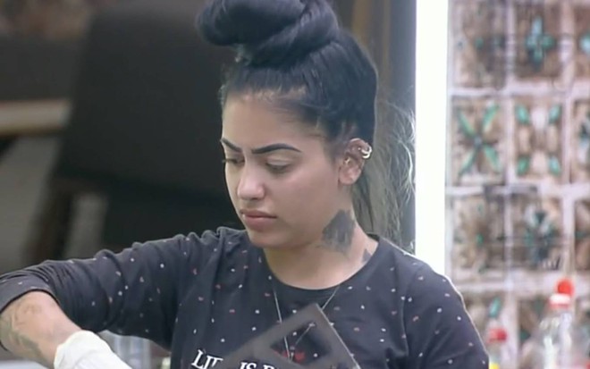 MC Mirella faz cara de nojo enquanto limpa a sede. A peoa está de cabelo preso, usa camiseta preta e luva branca nas mãos