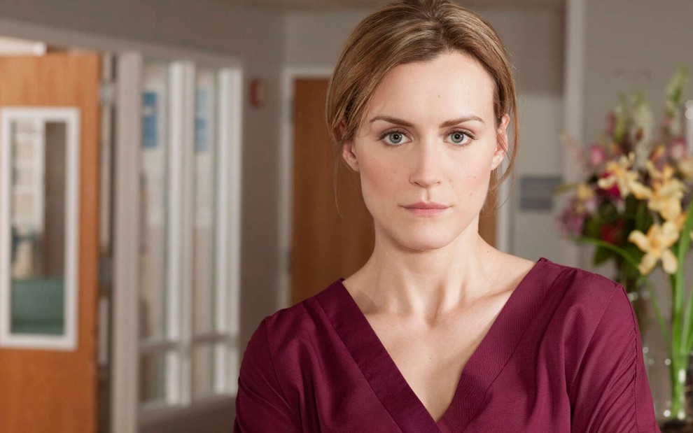 Em um colsutório médico, a loira Taylor Schilling veste um avental grená e faz cara série em imagem do drama Mercy