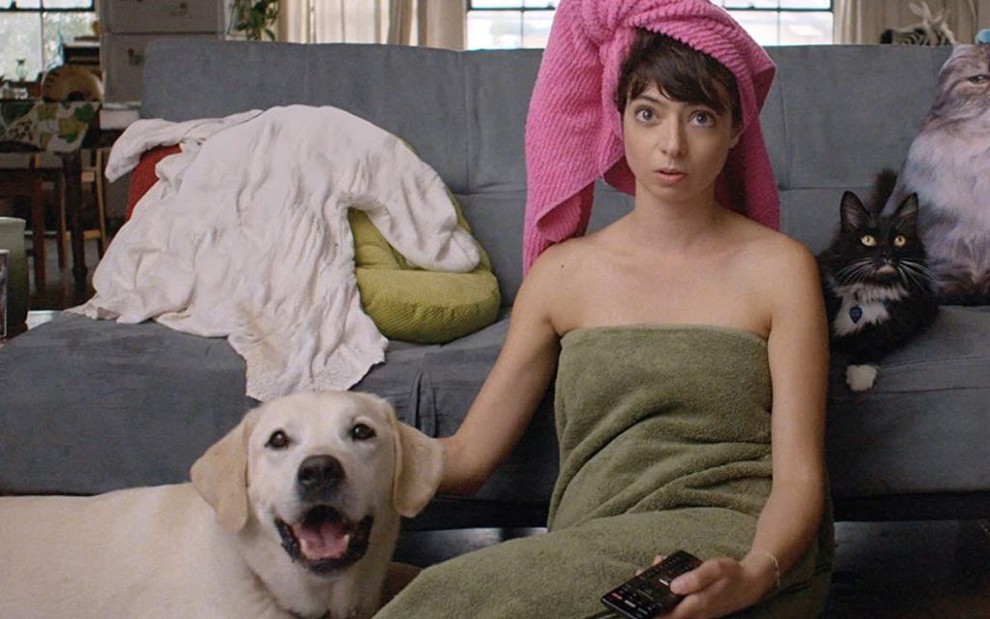 Kate Micucci de toalha enrolada no corpo e na cabeça assistindo TV com seus animais em cena do filme Meu Namorado É o Bicho