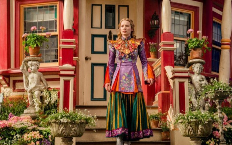 Mia Wasikowska como Alice em Alice Através do Espelho, com expressão séria, na frente de uma casa, usando um vestido longo nas cores laranja, lilás, verde e amarelo