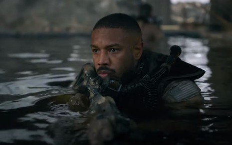 Michael B. Jordan como o personagem John Kelly aparece dentro da água, carregando uma metralhadora, com olhar atento em cena de Sem Remorso