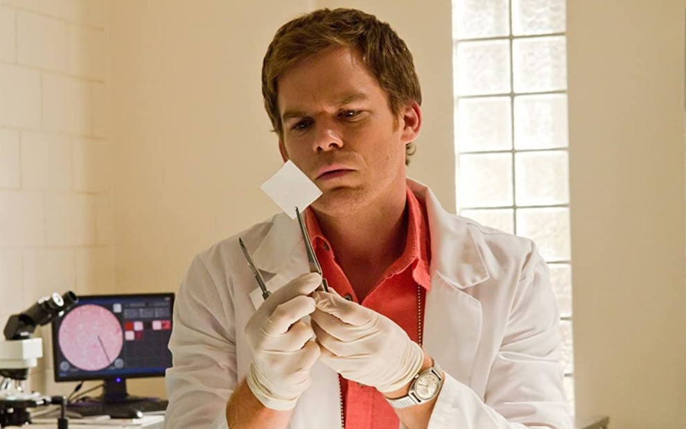 O ator Michael C. Hall, usando um avental branco, examina um pedaço de papel em cena de Dexter (2006-2013)