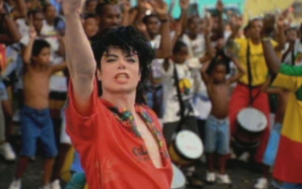 Com camisa vermelha e de gola bem aberta, Michael Jackson ergue o braço em posição de desafio; o grupo Olodum está atrás, durante gravação do clipe They Don't Care About Us em Salvador