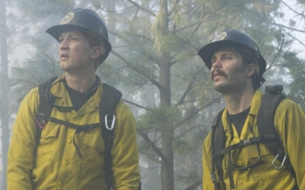 Em uma floresta cheia de fumaça, Miles Teller e Taylor Kitsch usam roupas amarelas e capacete e olham para o horizonte em cena de Homens de Coragem (2017)