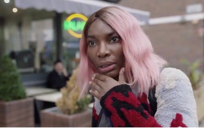 Arabella (Michaela Coel) de cabelo rosa e blusa branca e vermelha com expressão de espanto e mão esquerda no queixo