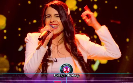 Cantora com cabelo preto comprido, blusa clara, segurando o microfone enquanto se apresenta no reality Vem Cantar!