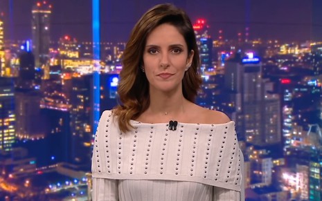 Monalisa Perrone com uma blusa tomara-que-caia clara, na bancada de um telejornal