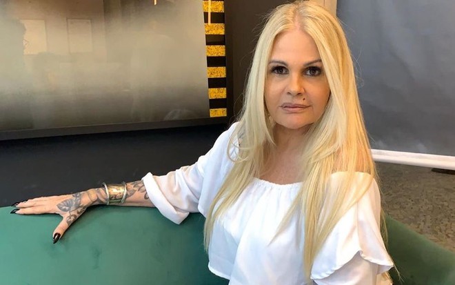 Monique Evans sentada em um sofá verde, de cabelo solto e blusa branca