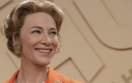 De vestido laranja e cabelo curto, a atriz Cate Blanchett solta um largo sorriso em cena da minissérie Mrs. America