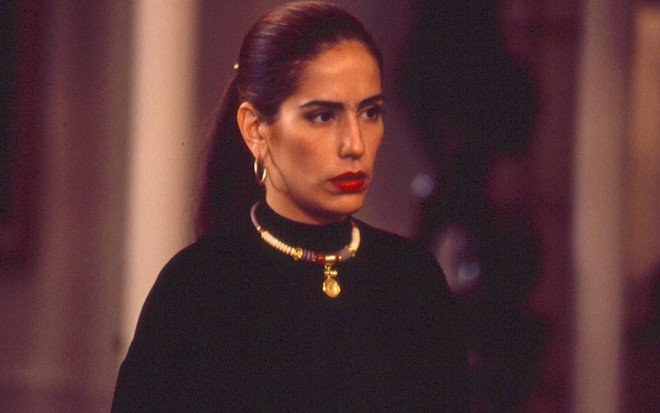 A atriz Gloria Pires com blusa preta, colar dourado, cabelo preso e expressão séria em cena como Raquel