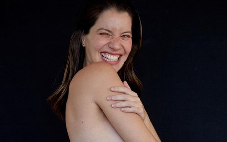 Nathalia Dill sem blusa, com mão no braço, de perfil; foto exibe apenas do peito para cima; ela sorri e pisca um olho
