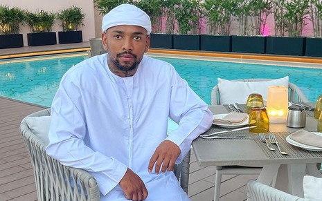 Nego do Borel usando roupa árabe, branca, sentado à beira da piscina de hotel de luxo